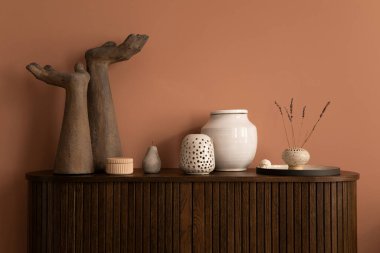Interior design of harmonized living room interior with wooden commode, modern hands sculpture, vase with dried flowers, beige casket, pink wall and personal accessories. Home decor. Template.