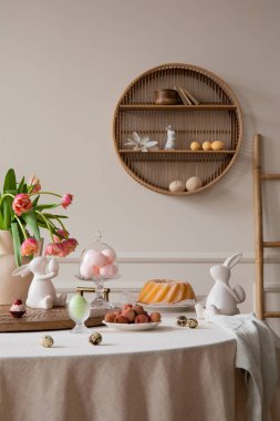 Interior design of easter dining room interior with round table, shelf on wall, colorful eggs, easter cake, hare sculptures, vase with tulips, lychee and personal accessories. Home decor. Template.