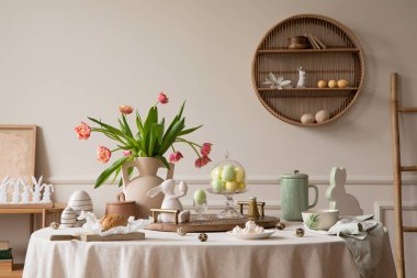 Creative composition of spring easter dining room interior with round table, vase with tulips, colorful eggs, pitcher, wooden trace, bread, hare sculpture and personal accessories. Home decor. Template. 