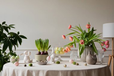 Interior design of easter dining room with colorful easter eggs, white hare sculptures, vase with tulips, plants, lamp, beige wall with stucco, gray hen and personal accessories. Home decor. Template.
