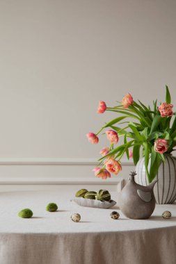 Minimalist composition of easter dining room interior with copy space, vase with tulips, colorful eggs, gray hen sculpture, beige wall with stucco and personal accessories. Home decor. Template. 