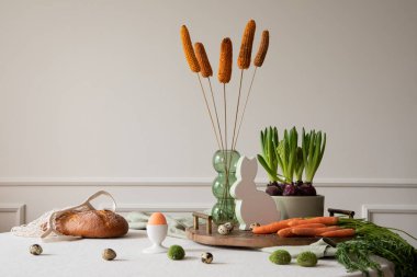 Creative composition of dinning room interior with round table, wooden trace, vase with dried flowers, orange carrot, hyacinth, hare sculpture, eggs and personal accessories. Home decor. Template.