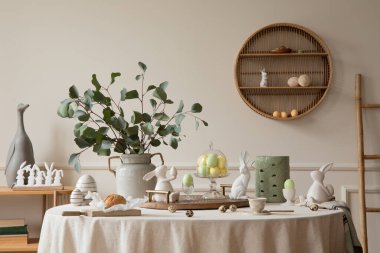 Warm and spring dining room interior with easter accessories, round table, vase with green leaves, cake, colorful eggs, rabbit sculpture and personal accessories. Home decor. Template. 