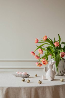 Minimalist composition of easter dining room interior with copy space, vase with tulips, colorful eggs, gray hen sculpture, beige wall with stucco and personal accessories. Home decor. Template. 