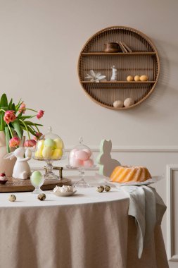 Interior design of easter dining room interior with round table, shelf on wall, colorful eggs, easter cake, hare sculptures, vase with tulips and personal accessories. Home decor. Template.