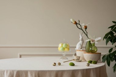 Minimalist composition of easter dining room interior with copy space, gray easter bunny sculpture, colorful eggs, vase with magnolia, beige wall with stucco and personal accessories. Home decor. Template.