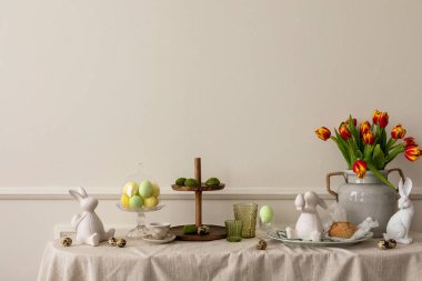 Creative composition of easter living room interior with copy space, vase with spring tulips, easter bunny sculpture, elegant cup, colorful easter eggs and personal accessories. Home decor. Template.