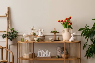 Warm and cozy composition of easter  living room interior with mock up poster frame, vase with tulips, hare sculpture, carrot, bowl, wooden sideboard and personal accessories. Home decor. Template. 