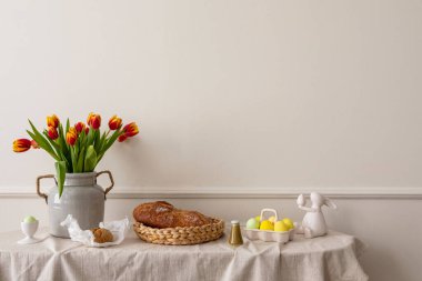 Minimalist composition of living room nterior with copy space, vase with tulips, bread in basket, colorful easter eggs, easter bunny sculpture and personal accessories. Home decor. Template.