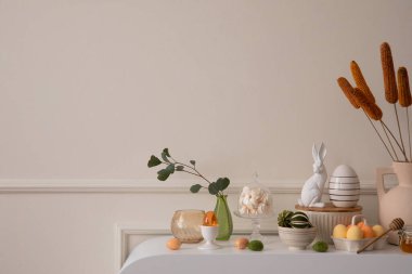 Minimalist composition of easter living room interior with copy space, easter bunny sculpture, colorful easter eggs, vase with leaves, wall with stucco and personal accessories. Home decor. Template.