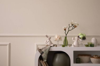 Minimalist composition of easter living room interior with copy space, easter bunny sculpture, easter eggs, vase with magnolia, wall with stucco and personal accessories. Home decor. Template.