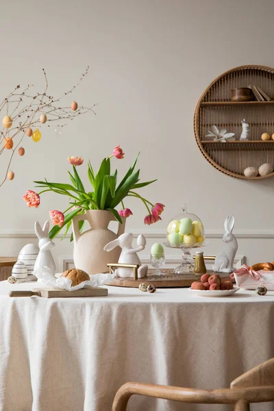 Creative composition of spring easter dining room interior with round table, vase with tulips, colorful eggs, wooden trace, bread, easter bunny, sculpture and personal accessories. Home decor Template