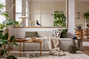 Open space interior with modular sofa, wooden coffee table, big window, patterned pillows, braided plaid, stylish lamp, beige coffee table, plants and personal accessories. Home decor. Template.