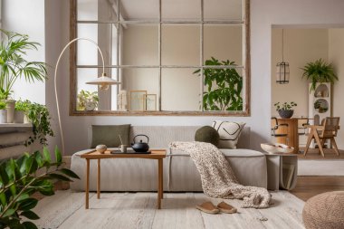 Warm and cozy interior with modular sofa, wooden coffee table, big window, patterned pillows, braided plaid, stylish lamp, wooden coffee table, plants and personal accessories. Home decor. Template.