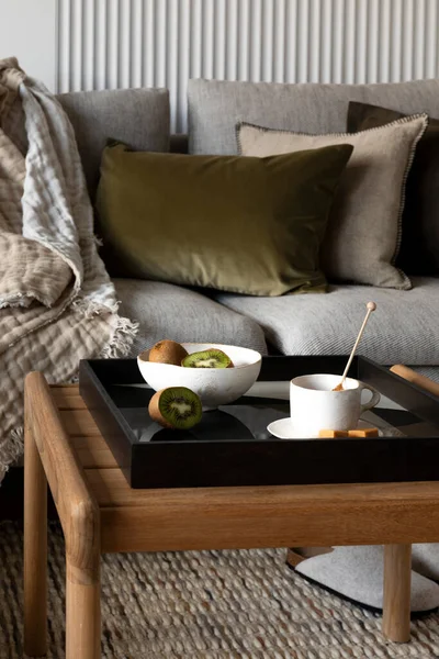 Creative composition of living room interior with wooden coffee table, bowl of kiwi, cup, simple black tray, gray sofa with pillows, plaid and personal accessories. Home decor. Template.