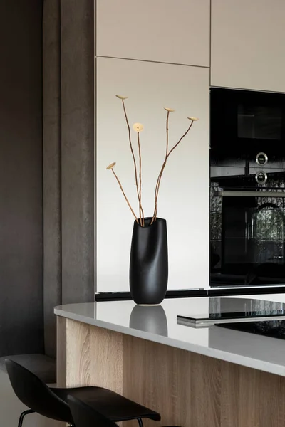 Modern composition of kitchen space with design kitchen island, design vase with dried flowers, black hookers, grey table, flowers, furnitures, big window and elegant personal accessories. Stylish home decor. Template.
