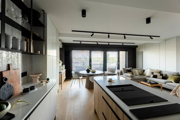 Modern composition of kitchen space with design kitchen island, black hookers, black induction hob, grey table, flowers, furnitures, big window and elegant personal accessories. Stylish home decor. Template.