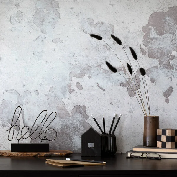 Creative composition of home office interior with black desk, ornaments on the wall, gray concrete wall, pencil holder and office accessories. Home decor. Template.