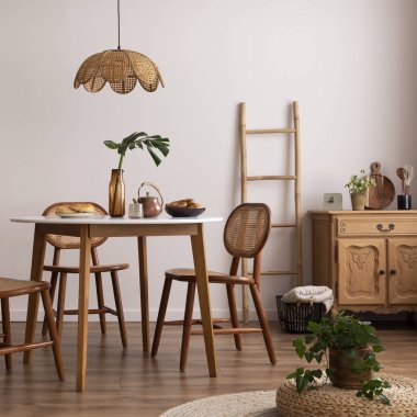 The stylish dining room with round table, rattan chair, wooden commode, poster and kitchen accessories. Beige wall with mock up poster. Home decor. Template. 