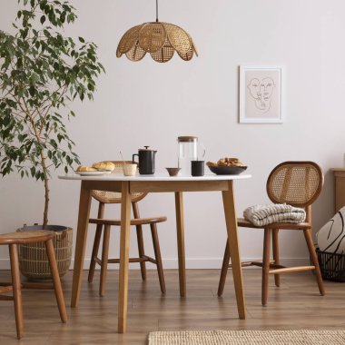 The stylish dining room with round table, rattan chair, wooden commode, poster and kitchen accessories. Beige wall with mock up poster. Home decor. Template. 