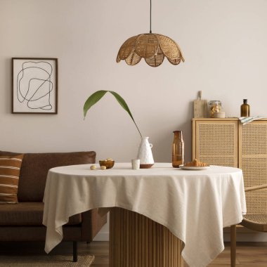 Boho and cozy interior of dining room with round wooden table, design chair, brown sofa, commode, mock up poster frame, pendant lamp, tea pot, decoration and elegant accessories. Stylish home decor. 