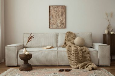 Interior design of living room with mock up poster frame, modern beige sofa, brown braided blanket, wooden coffee table, vase with dried flowers, round pillow, commode, vintage carpet and personal accessories. Home decor. Template.