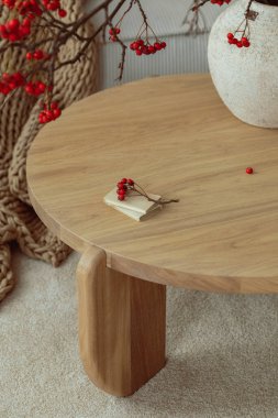 Minimalist composition of japandi living room interior with round wooden coffee table, stylish vase with rowan, simple beige carpet, braided plaid and personal accessories. Home decor. Template.