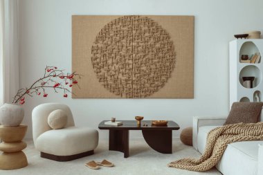 Japandi living room interior with mock up poster frame, modern black coffee table, rounded shapes armchair, beige sofa, vase with rowan, braided plaid and personal accessories. Home decor. Template.