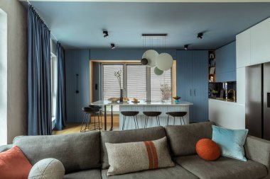 Interior design of kitchen interior with marble kitchen island, black chokers, blue kitchen furnitures, modern lamp, window, gray sofa, colorful pillows and personal accessories. Home decor. Template.