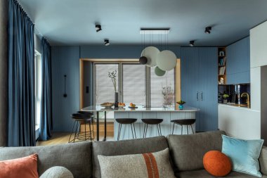 Interior design of kitchen interior with marble kitchen island, black chokers, blue kitchen furnitures, modern lamp, window, gray sofa, colorful pillows and personal accessories. Home decor. Template.