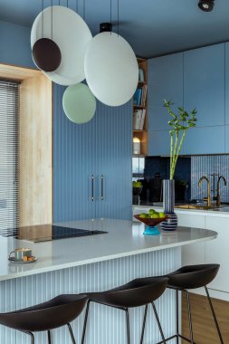 Interior design of kitchen interior with marble kitchen island, blue wall, black chokers, bowl with fruits, big window, cup, wooden floor, lamp on wall and personal accessories. Home decor. Template.