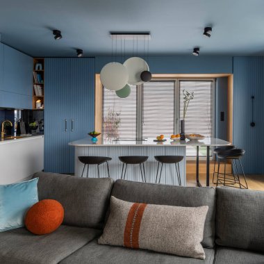 Interior design of kitchen interior with marble kitchen island, black chokers, blue kitchen furnitures, modern lamp, window, gray sofa, colorful pillows and personal accessories. Home decor. Template.