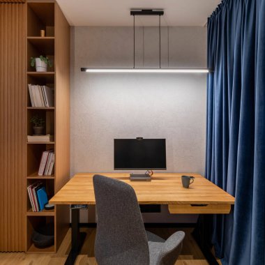 Interior design of office space with wooden desk, stylish gray armchair, navy curtain, modern lamp, computer, wooden bookcase, books, cup and personal accessories. Home decor. Template. 