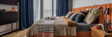 Warm and cozy bedroom interior with big orange bed, beige and grey bedclothes, blue curtain, tv, wooden night stand, black consola, round pillow, and personal accessories. Home decor. Template. 