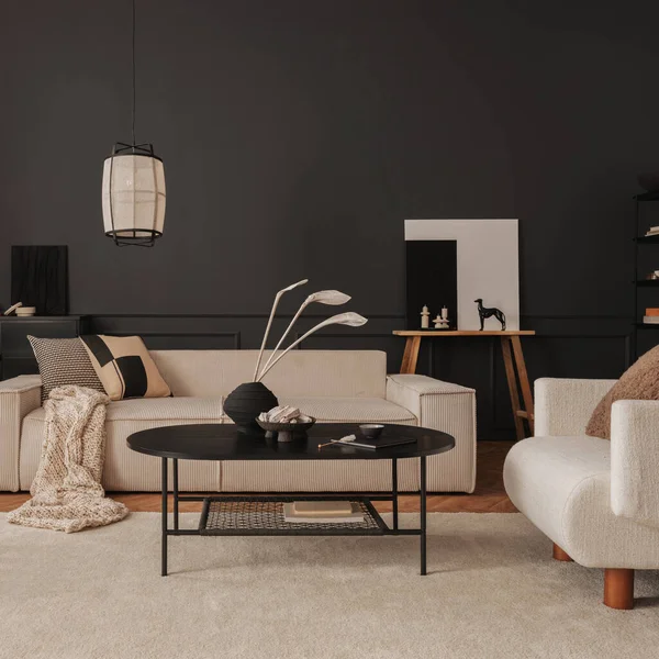Creative composition of living room interior with modular beige sofa, black coffee table, armchair, rug, black wall with stucco, vase with dried flowers and personal accessories. Home decor. Template.
