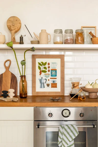 Cozy composition of kitchen interior with mock up poster frame, vase with flowers, white tile, wooden countertop, jar with spices, silver oven, vegetable and personal accessories. Home decor. Template
