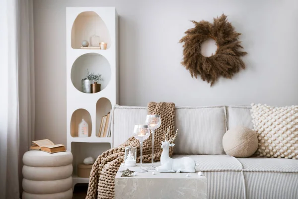 Amazing composition on white design shelf with christmas decoration, lights, gifts, lanterns, deer, candles, stars, round wreath white corduroy sofa and pouf. Santa claus is coming. Home decor. Template.