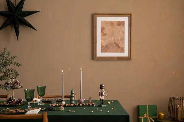 Aesthetic composition of christmas dining room interior with mock up poster frame, table, green tablecloth, candle with candlestick, brown wall, gifts, and personal accessories. Home decor. Template.