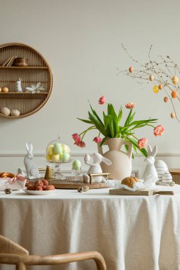 Creative composition of spring easter dining room interior with round table, vase with tulips, colorful eggs, wooden trace, bread, easter bunny, sculpture and personal accessories. Home decor Template