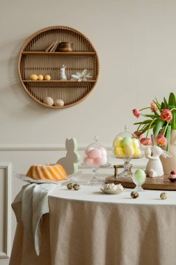 Interior design of easter dining room interior with round table, shelf on wall, colorful eggs, easter cake, hare sculptures, vase with tulips and personal accessories. Home decor. Template.