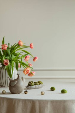 Minimalist composition of easter dining room interior with copy space, vase with tulips, colorful eggs, gray hen sculpture, beige wall with stucco and personal accessories. Home decor. Template.