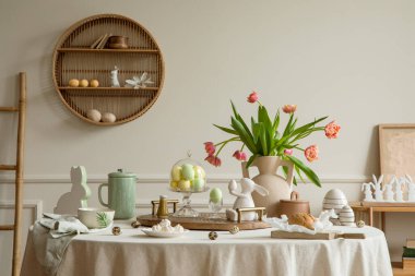 Creative composition of spring easter dining room interior with round table, vase with tulips, colorful eggs, pitcher, wooden trace, bread, hare sculpture and personal accessories. Home decor. Template.