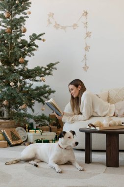 Noel süslemeleri olan şık bir oturma odası düzenlemesi. Genç bir kadın Noel ağacının yanında hediyeleri paketliyor. Noel yaklaşıyor. Ev dekorasyonu hoşmuş. Şablon.