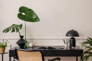 Siyah masa, rattan sandalye, renkli heykel, yapraklı vazo, raflar, kitaplar, sıva ve kişisel aksesuarlı duvar ile modern iş yeri kompozisyonu. Ev dekoru. Şablon. 