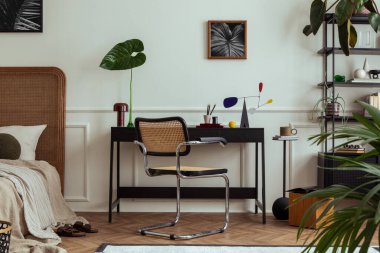 Poster çerçevesi, siyah masa, rahat yatak, yapraklı vazo, rattan sandalye, modern heykel, sıva ve kişisel aksesuarlı duvar olan odanın iç tasarımı. Ev dekoru. Şablon.