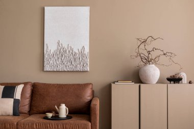 Poster çerçevesi, kahverengi kanepe, desenli yastık, bej sürahi, dallı vazo ve kişisel aksesuarları olan rahat oturma odasının estetik kompozisyonu. Ev dekoru. Şablon. 