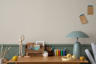 Ahşap masa, koltuk, nane lambası, kitaplar, şık heykeller, kalemler, sıva ve kişisel aksesuarlarla bej ve yeşil duvarlı samimi ofis iç mimarisi. Ev dekoru. Şablon.