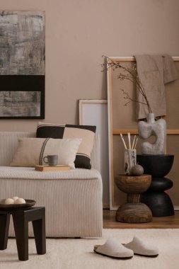 Poster çerçevesi, modüler kanepe, yastıklar, bej halı, ahşap tabure, gri terlikler, ahşap tezgah ve kişisel aksesuarlarla dekore edilmiş sıcak oturma odasının şık kompozisyonu. Ev dekoru. Şablon. 