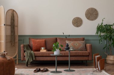 Kahverengi kanepe, turuncu yastık, ahşap bölme duvarı, örülmüş puf, yuvarlak masa, bitki ve kişisel aksesuarları olan sıcak bir oturma odası kompozisyonu. Ev dekoru. Şablon.