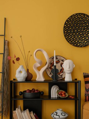 Siyah konsol, şık heykel, kurumuş çiçekler, siyah merdiven, renkli sepet, yastık ve kişisel aksesuarlarla dolu sarı oturma odasının yaratıcı kompozisyonu. Ev dekoru. Şablon